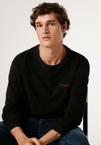 Črna majica z dolgimi rokavi z rdečim vezenim napisom na prsih. Prijeten kroj z okroglim izrezom in mehkim materialom.