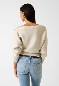 Kremowy, prążkowany sweter z szerokim dekoltem odsłaniającym ramiona, bufiastymi rękawami i ozdobnymi guzikami z tyłu, zestawiony z jasnoniebieskimi dżinsami.