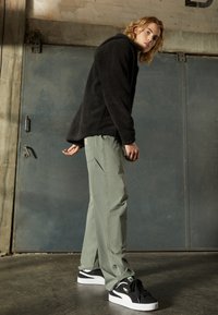 Veste en polaire noire en peluche avec capuche, associée à un pantalon cargo vert olive clair et des baskets noires avec des accents blancs.