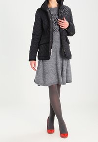Black quilted jacket with a high collar, paired with a gray patterned dress. Model wears gray tights and red high heels.