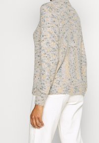 Persoon in een lichtgrijze en gele gespikkelde gebreide trui met lange mouwen en witte broek, gezien van opzij en van achteren.