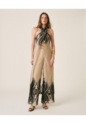 Mujer con mono de pierna ancha beige con estampados florales negros, escote halter, pulsera de oro ancha y tacones abiertos negros, sobre fondo neutro.