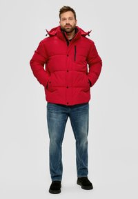 Rote gepolsterte Winterjacke mit Kapuze, ausgestattet mit einer Reißverschlusstasche auf der Brust und Knopfverschluss. Kombiniert mit blauen Jeans und schwarzen Schuhen.