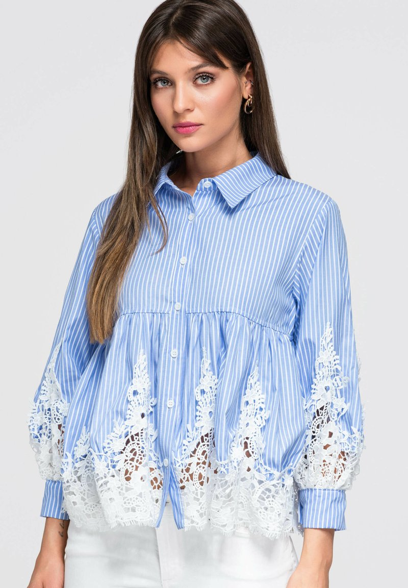 Femme portant un chemisier à rayures bleues et blanches avec des boutons et des détails en dentelle blanche à l'ourlet et aux manches, posant devant un fond uni.