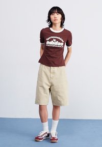 Brown short-sleeve t-shirt with white accents and graphic print, paired with beige shorts, white socks, and maroon athletic shoes.