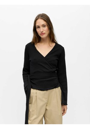 Kvinde med langt hår iført sort vævet sweater med sidelukning og beige plisserede bukser, stående foran en ensfarvet hvid baggrund.