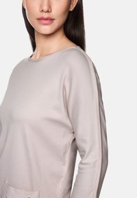 Helle beige Langarmbluse mit Rundhalsausschnitt, die über eine gerippte Textur und Details an den Seitennähten verfügt, einschließlich einer Tasche auf der Vorderseite.