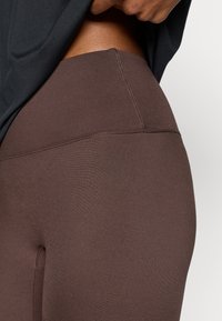 Καφέ κολάν από λείο, ελαστικό ύφασμα. Διαθέτει ψηλή μέση και επίπεδες ραφές χωρίς ορατά σχέδια ή μεταλλικά στοιχεία.