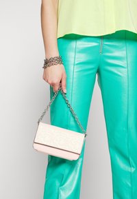 Mała różowa torebka z tłoczonym wzorem, srebrnym łańcuszkowym paskiem, o prostokątnym kształcie, w zestawie z zielonymi skórzanymi spodniami i żółtą bluzką.