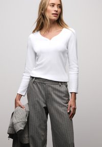 Witte top met lange mouwen en een V-hals met drie knopen, gecombineerd met grijze gestreepte broek en een grijze jas over één arm gedrapeerd.