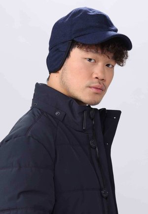 Navy blue wool cap with ear flaps, curved brim, and flat crown, worn with a black padded jacket featuring a high collar and snap fasteners.