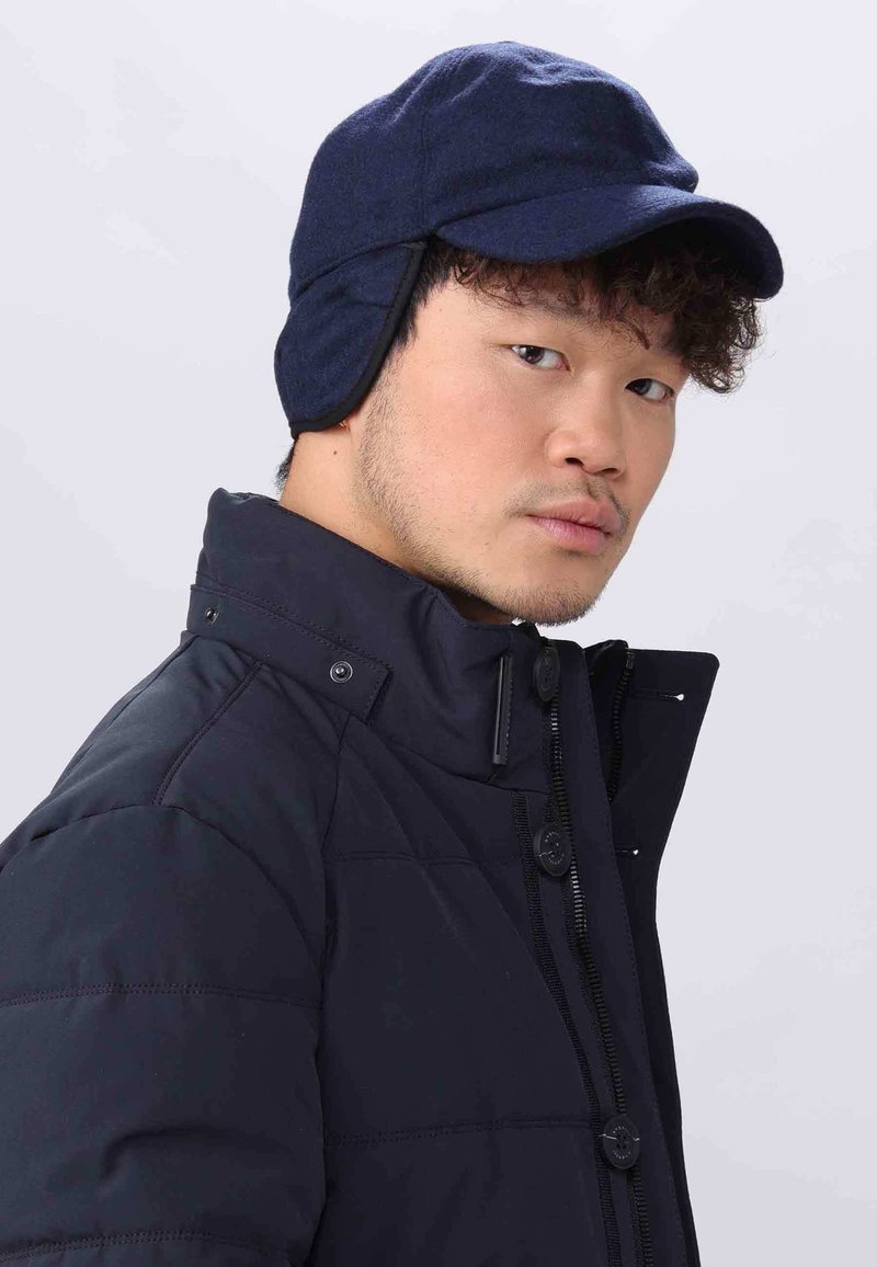 Marineblaues Wollmütze mit Ohrenschutz, gebogenem Schirm und flachem Oberteil, getragen mit einer schwarzen gepolsterten Jacke mit hohem Kragen und Druckknöpfen.