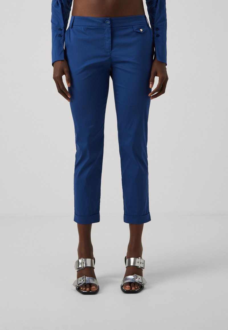 Pantalons bleus cropped en tissu lisse avec un design ajusté, dotés d'une fermeture à bouton et de poches retournées, accompagnés de sandales à accents argentés.