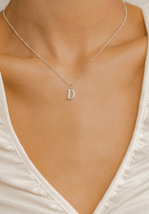 Silver necklace with a textured chain and a sparkling letter "D" pendant, set against smooth skin and a white top.