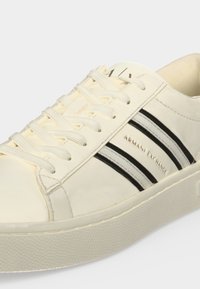 Κρεμ χρώματος αθλητικά παπούτσια Armani Exchange με λευκά κορδόνια και δύο ασπρόμαυρες ριγέ λεπτομέρειες στην πλευρά.