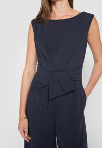 Marineblå jumpsuit med detaljer på skuldrene, med et knyttet taljebånd og vidde i benene. Fremstillet af glat stof med en let glans.