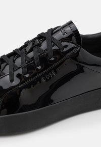 Baskets en cuir verni noir avec semelle en caoutchouc noir texturé, présentant des lacets noirs et un logo « BOSS » embossé sur le côté.