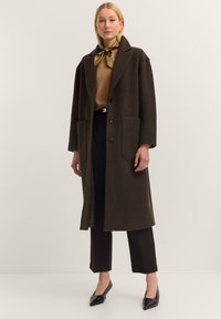 Dunkelbrauner Wollmantel mit Reverskragen, oversized Schnitt, zwei vorderen Taschen und Knopfverschluss, getragen über einem beigen Oberteil und schwarzen Hosen.