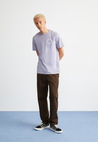 Camiseta de algodón lavanda con un diseño circular impreso en el pecho, combinada con pantalones rectos marrones y zapatillas a cuadros en blanco y negro.
