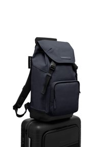 Un sac à dos bleu marine avec un design à rabat, des bretelles réglables, une poche avant et un tissu texturé, posé sur une valise noire.