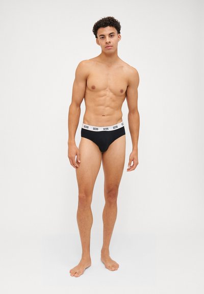 Calzoncillos cortos negros con una cinturilla elástica blanca que presenta el logo de la marca, exhibidos en un modelo de pie con un fondo neutro.