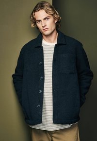 Schwarze Fleecejacke mit Knopfverschluss, versehen mit einer Brusttasche. Getragen über einem weißen und marineblauen gestreiften Hemd, kombiniert mit khakifarbenen Hosen.