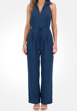 Granatowy kombinezon bez rękawów z kołnierzem i ściągaczem w talii, noszony przez kobietę stojącą, do którego dobrała białe sandały na obcasie z odkrytymi palcami oraz złotą biżuterię.