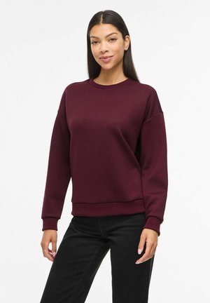 Femeie cu păr lung și închis la culoare, purtând un pulover simplu bordo cu mâneci lungi și pantaloni negri, stând în picioare pe un fundal alb.