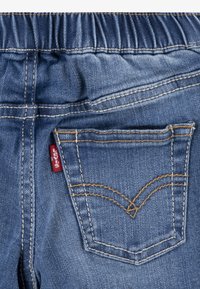 Modri kavbojski jeans s elastikim pasom, ki vključujejo zadnji žep z rumenim šivom in majhno rdečo oznako blagovne znamke. Gladka tekstura.