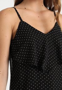 Černá top s puntíky, tenké ramínka, volánová vrstva vpředu a lehký materiál s malými bílými tečkami.