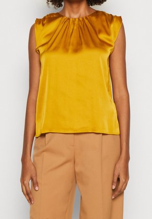 Femme portant un chemisier sans manches brillant jaune moutarde associé à un pantalon sur mesure marron clair sur un fond clair uni.