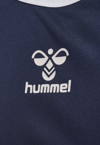 Białe stylizowane logo pszczoły oraz napis „hummel” nadrukowane na granatowej tkaninie, prawdopodobnie część odzieży sportowej lub treningowej.