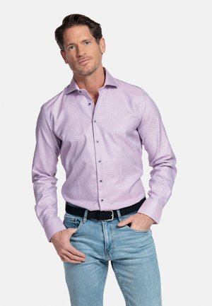 Man met een lichtpaars geruit overhemd met lange mouwen en een lichtblauwe spijkerbroek, staand met handen in de zakken tegen een witte achtergrond.