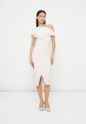 Kobieta stojąca, ubrana w białą, jednobarkową sukienkę midi z asymetrycznym dołem oraz dopasowane szpilki na spiczastym nosku na jednolitym białym tle.