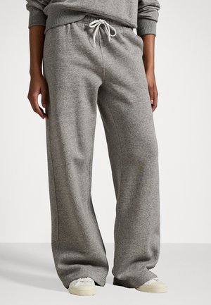 Henkilö, jolla on päällään väljät harmaat collegehousut valkoisella nyörillä ja valkoiset lenkkarit, seisoo yksinkertaisen vaalean taustan edessä.