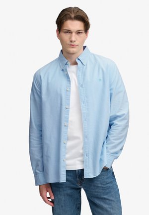 Ung mand iført en lyseblå skjorte med knapper over en hvid t-shirt og blå jeans, stående med den ene hånd i lommen.