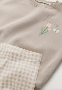 Bež pulover z vezenim cvetličnim motivom in napisom "Les Fleurs", skupaj z bež in belim karo tkanjem. Mehak bombažni material.