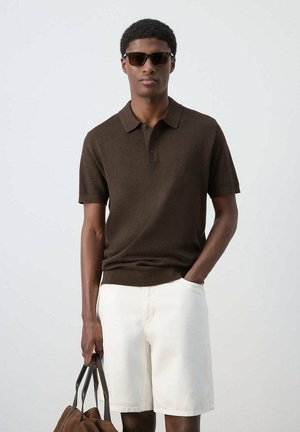 Hombre con gafas de sol, camiseta tipo polo de color marrón oscuro, pantalones cortos blancos, sostiene una bolsa marrón en una mano, la otra mano en el bolsillo, de pie frente a un fondo liso.