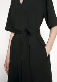 Robe noire avec ceinture, dotée d'un col en V, de manches courtes et de poches. Confectionnée dans un tissu lisse à la texture légère.