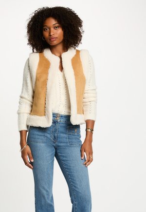 Donna con capelli ricci che indossa un maglione bianco di maglia, un gilet di shearling color beige, jeans blu a vita alta e bracciali e anelli d'oro.