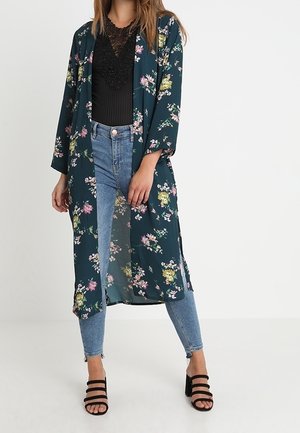 Woman wearing a dark floral long kimono, black lace top, light blue skinny jeans, and black strappy block heel sandals, standing on white floor.