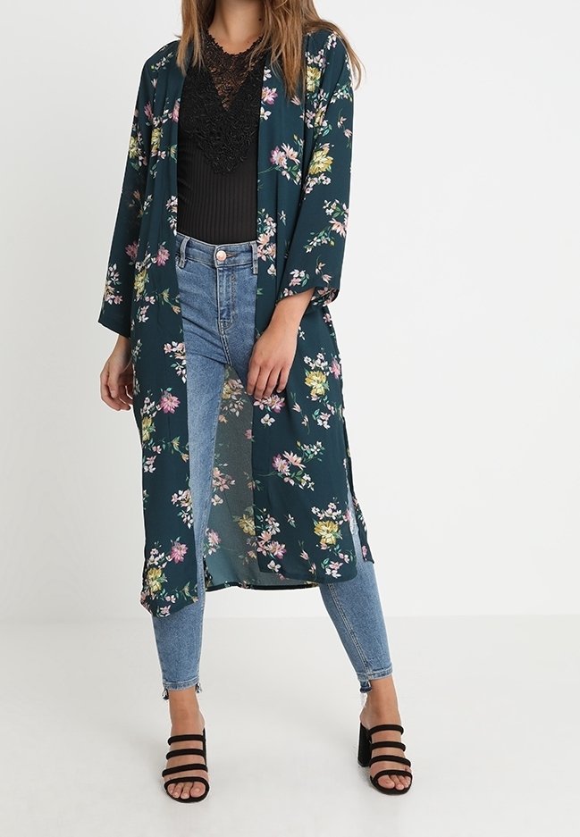 Femme portant un long kimono foncé à motif floral, un top en dentelle noire, un jean skinny bleu clair et des sandales noires à talons blocs et brides, debout sur un sol blanc.