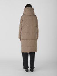 Doudoune marron clair, avec un col montant, un matelassage horizontal et un design mi-long. Assortie à un jean noir à jambes larges et des bottes noires.