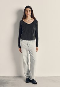 Ženska stoji ob nevtralni steni, oblečena v temnosivi dolg rokav, svetlo modre kavbojke in črne loaferje, obrnjena naravnost naprej z rokami sproščeno ob telesu.