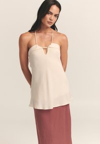 Haut sans manches beige clair avec une découpe en trou de serrure et un détail en perles en bois au niveau de l'encolure, associé à une jupe rose fluide. Texture lisse.