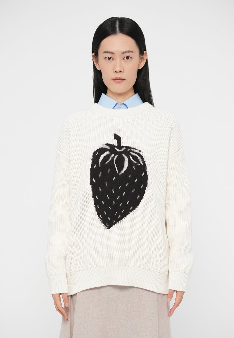 Camisola branca tricotada com um grande desenho de morango preto. Apresenta punhos e bainha canelados, ajuste relaxado e um decote aberto sobre uma camisa azul clara.