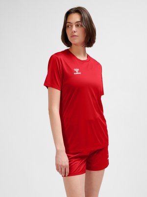 Vrouw die een rood sport-T-shirt en bijpassende korte broek draagt, staat met ontspannen armen en kijkt opzij tegen een effen achtergrond.