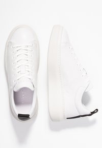 Białe skórzane trampki z okrągłymi noskami, płaskimi sznurówkami, teksturowanymi podeszwami i czarnym akcentem z tyłu. Minimalistyczny design bez widocznych logo.