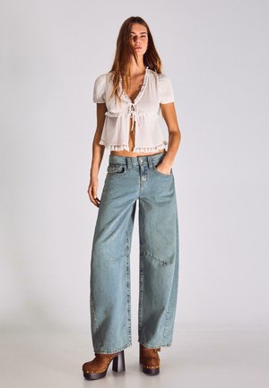 Kobieta stojąca z jedną ręką w kieszeni, ubrana w białą, krótką bluzkę z falbankami, szerokie dżinsy i brązowe buty na platformie.