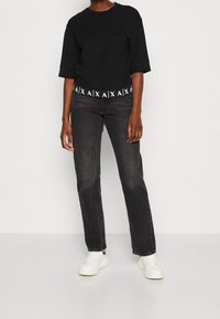 Černé zkrácené tričko s širokými rukávy a bílým logem, spárované s rovnými tmavými džíny a bílými teniskami na neutrálním pozadí.